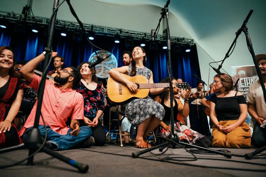 a group of people sitting on stage performing. one woman is holding a guitar.