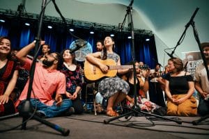 a group of people sitting on stage performing. one woman is holding a guitar.